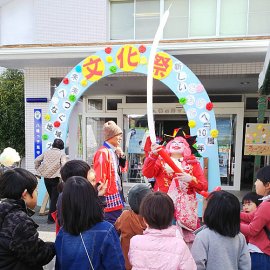 子ども達に囲まれる中細長い風船を膨らませるバルーンクラウン